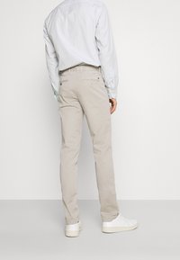 Beige Baumwoll-Chinos mit geradem Schnitt, ausgestattet mit Gesäßtaschen und einem dezenten Logo-Detail. Kombiniert mit einem hell gestreiften Hemd mit Knöpfen und weißen Sneakern.