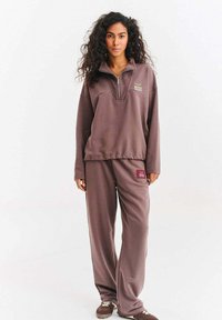 Kobieta stoi ubrana w dopasowany lawendowy sweter i spodnie z logo "Naoko Studio", do tego brązowe tenisówki, na jednolitym tle.