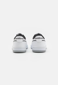 Bílé sportovní boty s černými akcenty, hladký syntetický materiál a strukturovaný podpatek. Vlastnosti zahrnují logo Reebok na zadní straně a nízký profil.