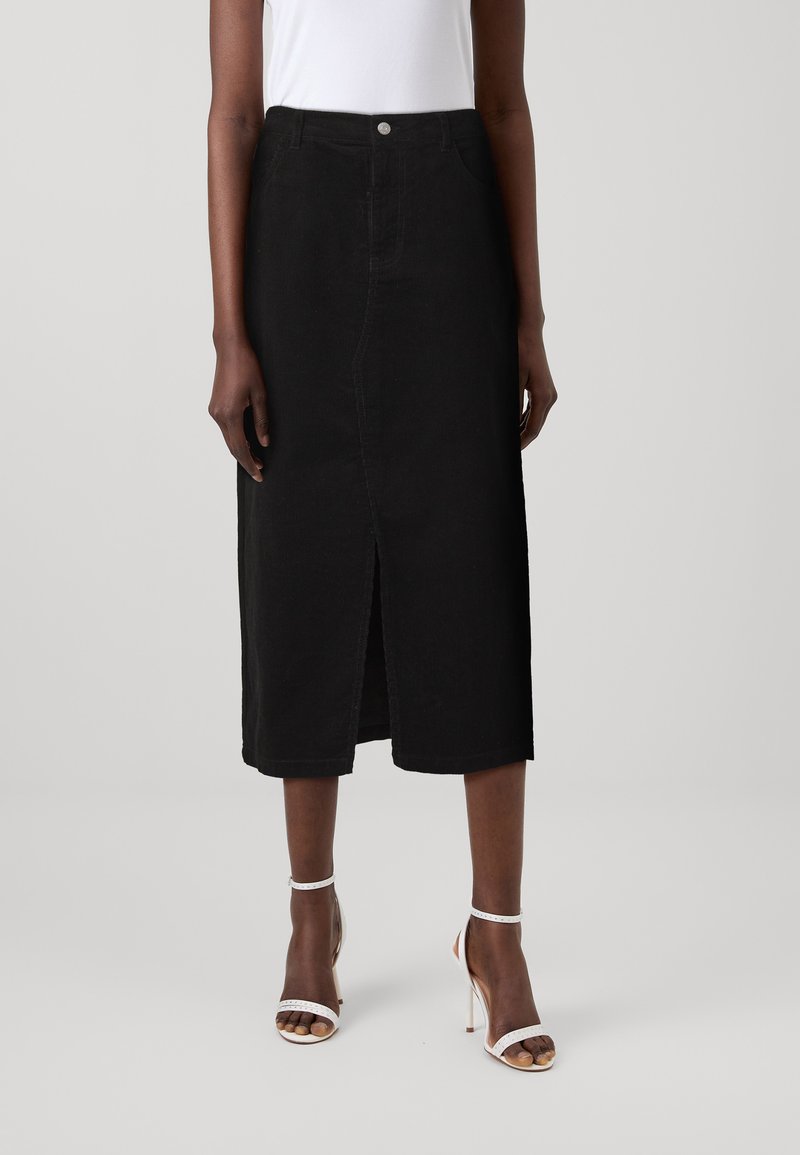 Jupe midi en denim noir avec une fente frontale, présentant une taille ajustée, des passants de ceinture et des poches latérales. Portée avec des sandales à talons blanches.