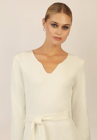 Cremefarbener Strickpullover mit V-Ausschnitt und langen Ärmeln, der mit einem gebundenen Taillengürtel akzentuiert ist. Weicher Stoff, minimalistisches Design.