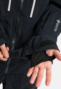 Schwarze Gore-Tex-Jacke mit einem Frontreißverschluss, verstellbaren Säumen mit Klettverschluss und einer gestrickten Innenschicht, die sich von den Ärmeln erstreckt.