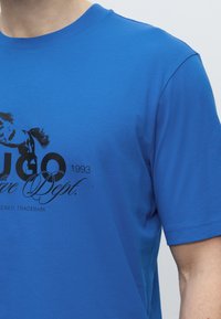 Μπλε μπλουζάκι με μαύρο κείμενο "HUGO 1993 Active Dept." και μαύρο γραφικό με πρόσωπο και μαλλιά ατόμου στο στήθος.