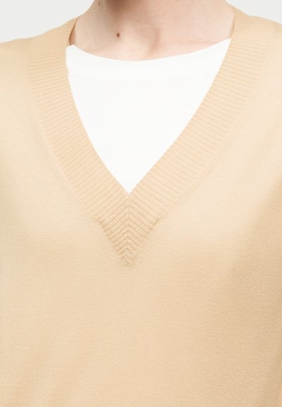 Pull beige à col en V superposé sur un t-shirt blanc à col rond, montrant l'encolure et le haut de la poitrine d'une personne.