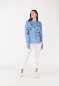Blauw gestreepte lange mouwen shirt met bloemembroidery detail aan de voorkant, gecombineerd met witte getailleerde broek en sneakers. Effen achtergrond.