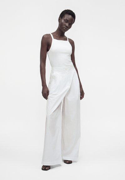 Femme debout portant un haut blanc sans manches à encolure carrée et un pantalon blanc large taille haute avec des sandales noires à bouts ouverts.