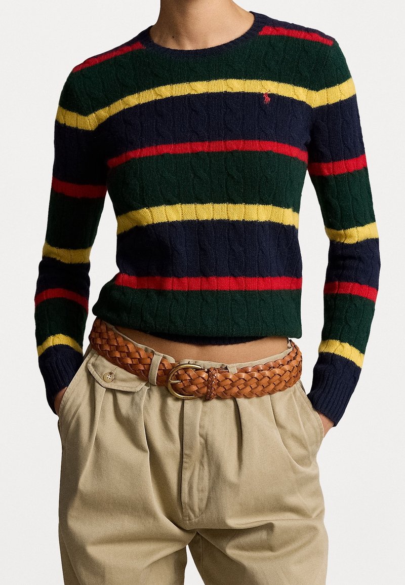 Personne portant un pull en maille torsadée rayé bleu marine, vert, rouge et jaune, rentré dans un pantalon beige taille haute avec une ceinture en cuir brun tressé.