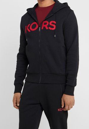 Homme portant un sweat à capuche noir zippé et un pantalon avec le texte rouge "KORS", associé à une chemise bordeaux en dessous.