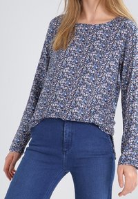 Woman wearing a long-sleeve navy floral top and high-waisted blue jeans, with loose blond hair visible on one side.