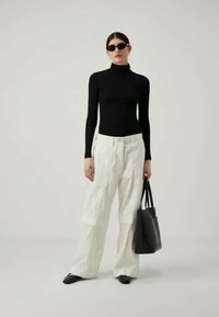 Woman wearing black turtleneck, white wide-leg pants, black shoes, sunglasses, holding black tote bag, standing against plain background.