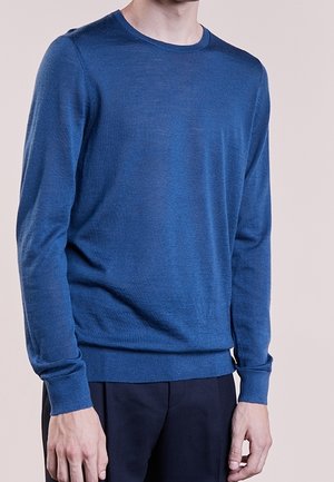 Mann trägt einen schlichten blauen Pullover mit Rundhalsausschnitt und langen Ärmeln sowie dunkle Hosen vor neutralem Hintergrund.