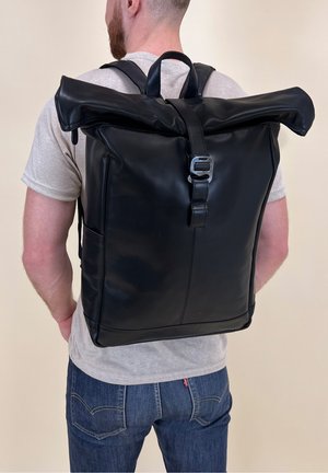 Homme portant un t-shirt gris clair et un jean bleu, portant un grand sac à dos en cuir noir à rabat roulé avec boucle en métal dans le dos.