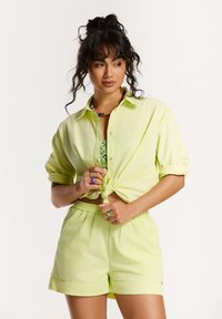 Vrouw met krullend donker haar die een knoop in haar lichtgroene overhemdbloes maakt, gekleed in bijpassende shorts en kleurrijke ringen en een ketting, terwijl ze zijwaarts kijkt.