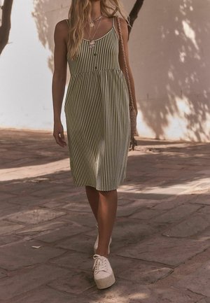 Frau, die auf Pflastersteinen geht und ein ärmelloses, grün-weiß gestreiftes Kleid, weiße Turnschuhe, mehrere Ketten und eine gewebte Umhängetasche trägt.