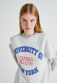 Jovem mulher com cabelo longo e ruivo, usando brincos de argola dourados e um sweatshirt cinza com o texto "Universidade de Nova Iorque NYC".