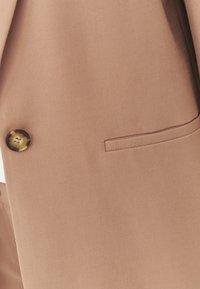 Gros plan sur le devant d'un blazer en tissu marron avec un bouton en écaille et un détail de poche passepoilée sur le côté droit.