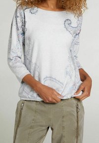 Sudadera gris claro con mangas tres cuartos, con diseño de cachemira colorido y adornos. Combinada con pantalones verde oliva con cremallera.