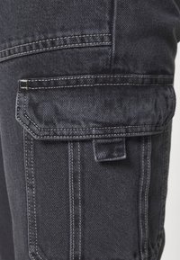 Pantalones cargo de mezclilla oscura con dos bolsillos laterales, costuras visibles y una tela texturizada. Presenta una solapa de bolsillo plegada con cierre a presión.