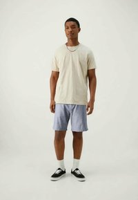 Lys beige t-skjorte, grå-blå shorts, svarte joggesko med hvite såler, og hvite sokker. Enkelt, avslappet antrekk vist på en person som står.