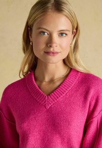 Pulover cu decolteu în V, de culoare roz strălucitor. Fabricat dintr-un material moale, texturat, cu accente ribbrate pe guler. Colier simplu din aur ca accesoriu.