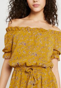 Off-shoulder blus i senapsgul, med en blommig print i lila och vitt, volangkrage och knytdetalj i midjan.