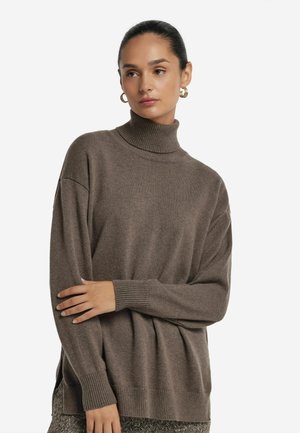 Kvinna i brun polotröja och mönstrade byxor, med armarna i kors och gyllene hoops örhängen, som ser lite åt sidan.