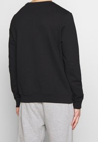 Felpa nera in misto cotone, con maniche lunghe, scollatura tonda e polsini a coste. Indossata con pantaloni della tuta grigi chiari.