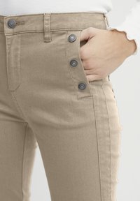 Pantalons beiges taille haute avec une texture lisse et cinq accents de boutons sur le côté. Comprend une poche avec un détail de couture subtil.