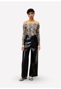 Vrouw met kort krullend haar, gekleed in een off-shoulder bloezen top, zwarte wijde leren broek en zilveren schoenen, staand tegen een witte achtergrond.