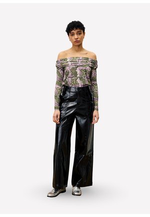 Femme aux cheveux courts et bouclés, portant un haut floral décolleté, un pantalon en cuir noir à jambes larges et des chaussures argentées, se tenant devant un fond blanc.