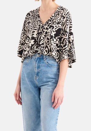 Frau trägt eine locker sitzende Bluse mit schwarz-weiß abstraktem Muster, Ellbogen langen Ärmeln und hoch taillierten hellblauen Jeans.