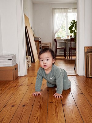 Un tout-petit vêtu de gris rampe sur un parquet en direction de la caméra. À l’arrière-plan, on aperçoit une salle à manger lumineuse et des plantes d’intérieur.