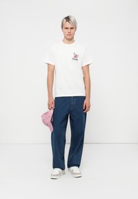 White cotton t-shirt with graphic print, loose blue denim jeans, holding a pink baseball cap, and wearing white sneakers with grey accents.