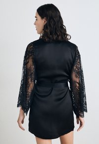 Robe di raso nero con maniche lunghe realizzate in preziosa pizzo nero, vita legata e una texture liscia, con un taglio che arriva fino al ginocchio.
