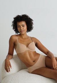 Beige BH og matchende truse laget av glatt stoff. BH-en har bøyler og tynne stropper. Modell sitter på en teksturert, lysfarget stol.