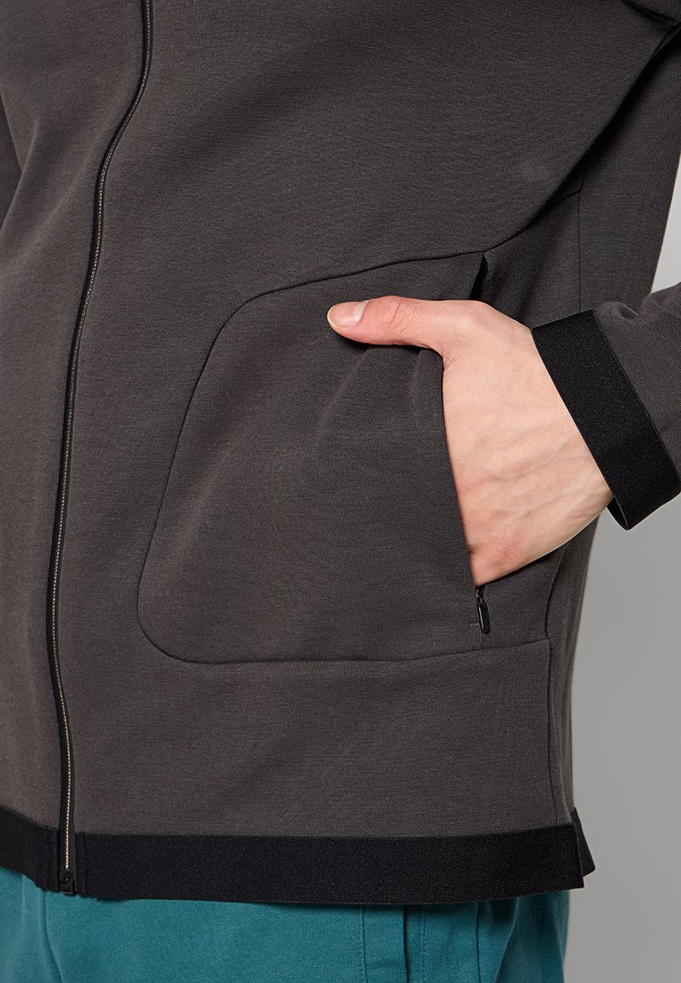 Giacca grigia scura realizzata in tessuto morbido, con una zip frontale prominente, ampio taschino laterale e rifiniture nere sui polsini e sull'orlo.