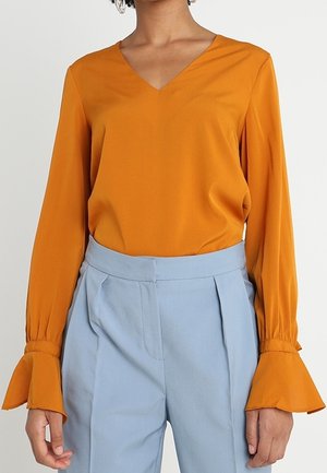 Blouse à manches longues en tissu moutarde avec décolleté en V et poignets évasés, associée à un pantalon taille haute bleu clair avec plis.