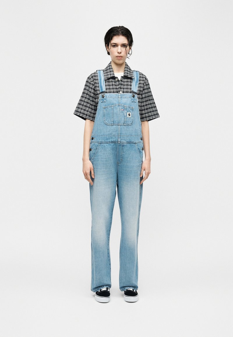 Personne debout devant un fond blanc, portant une salopette en jean bleu clair, une chemise à manches courtes à carreaux noirs et blancs, et des baskets noires.