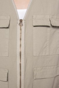 Beige utility vest with a zip front, two large chest pockets, and a textured fabric. Features a smooth zipper with a bronze finish.