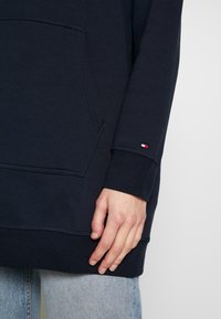Sudadera con capucha de color navy, con un bolsillo frontal, mangas acampanadas y un pequeño parche de logo en la manga. Textura de tela suave y corte relajado.