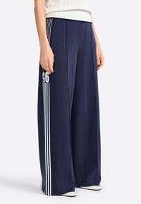 Pantalons larges bleu marine en tissu doux, avec des rayures verticales blanches sur les côtés et un détail de logo imprimé.