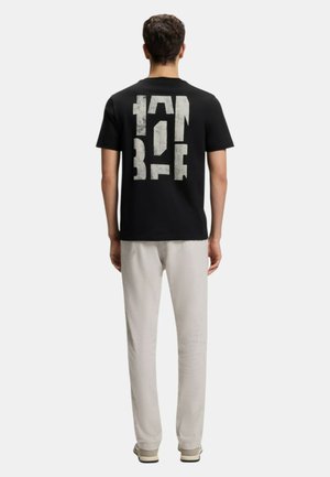 Homme portant un T-shirt noir à manches courtes avec un grand texte abstrait vertical blanc dans le dos, un pantalon beige et des baskets beiges, debout de dos.