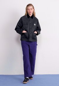 Schwarze, knöpfbare Jacke mit Kragen und aufgesetzten Taschen, kombiniert mit lila Hosen und schwarzen Schuhen, vor einem schlichten Hintergrund.