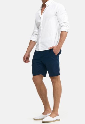 Weißes Leinenhemd mit Knöpfen, marineblaue Shorts und weiße Espadrilles. Das Hemd hat einen klassischen Kragen, während die Shorts seitliche Taschen haben.