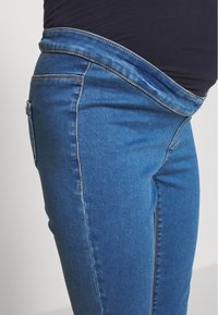 Jeans de maternidad de mezclilla elástica en azul medio. Con una cinturilla alta y diseño tradicional de cinco bolsillos. Textura suave y forma ajustada.