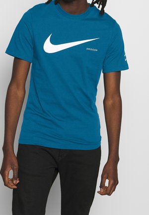 T-shirt turquoise à manches courtes en coton avec un grand logo Swoosh blanc de Nike et le texte "SWOOSH" sur le devant. Pantalon noir porté avec.
