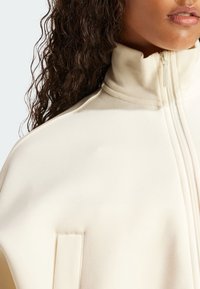 Krämlfärgad zip-up-jacka med hög krage, gjord av texturerat tyg. Har en sidoficka och synliga sömnadsdetaljer.