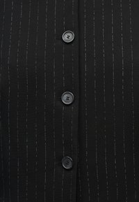 Μαύρο ύφασμα με λεπτές λευκές κάθετες ρίγες, με τρία στρογγυλά μαύρα κουμπιά ραμμένα σε κάθετη γραμμή.