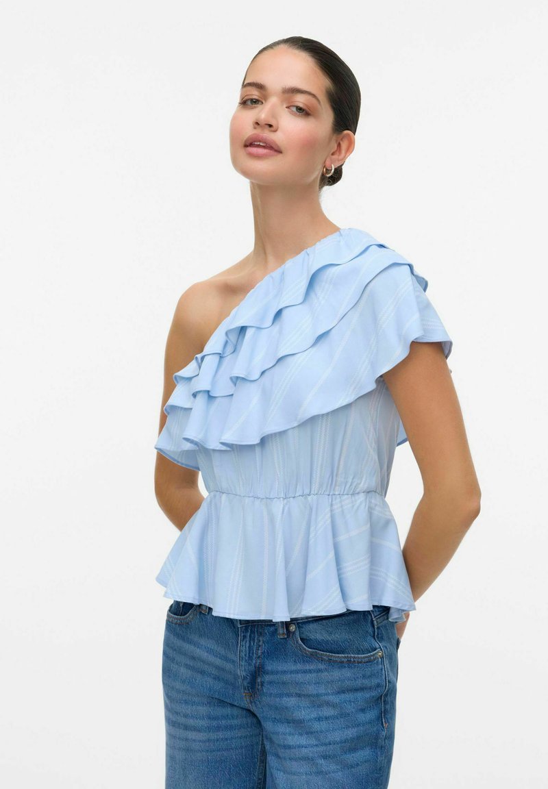 Haut bleu clair à une épaule avec un design en volants, présentant des rayures verticales et une taille peplum. Porté avec un jean bleu.