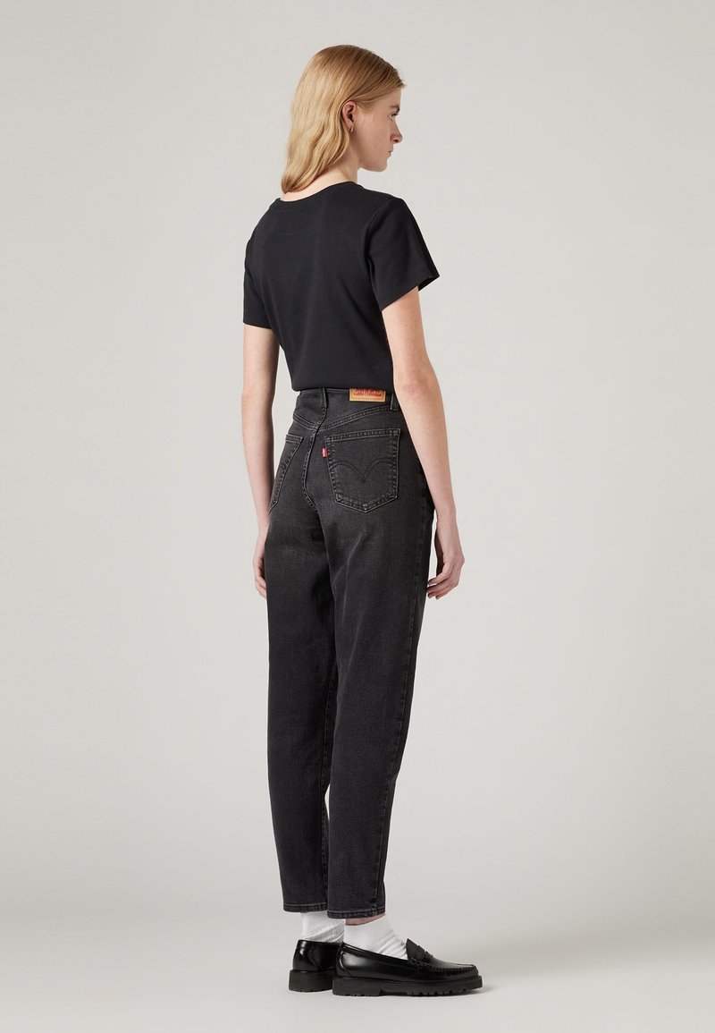 Kobieta z blond włosami nosi czarną koszulę z krótkim rękawem, czarne spodnie z wysokim stanem, białe skarpetki i czarne mokasyny, stojąc na jednolitym tle.